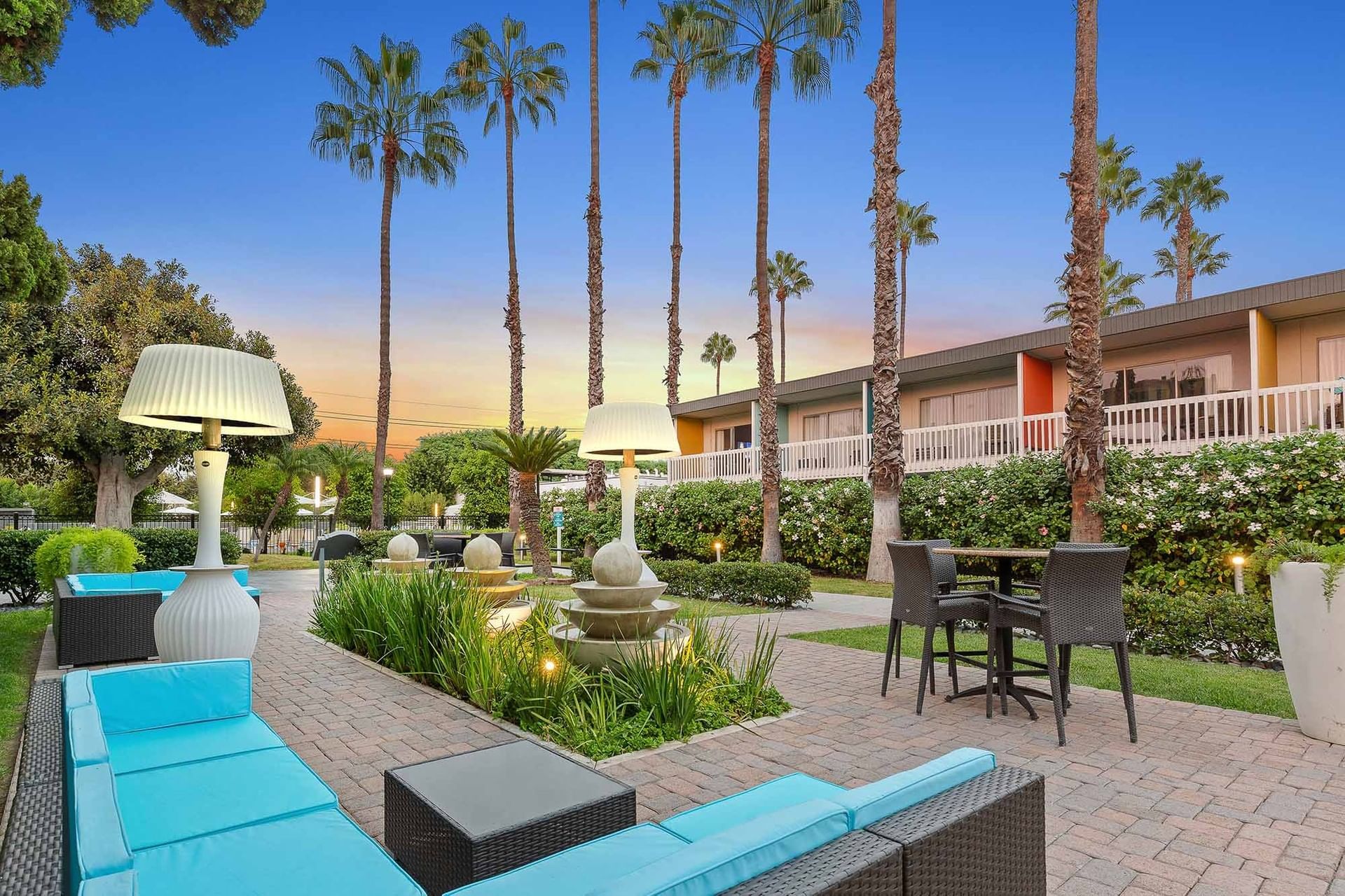 A serene outdoor seating area with a fountain, palm trees, and outdoor furniture at sunset.