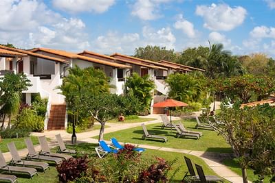 Row of white resort villas with red roofs beside a grassy lawn and lounge chairs under trees at Sugar Cane Club Hotel & Spa