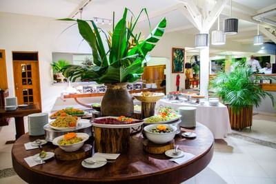 Round wooden table with buffet dishes, salads, & a clay vase of tropical leaves in Acajou Restaurant at Hotel Montana Haiti