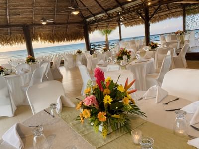 Open-air beachfront dining pavilion with thatched roof, white-draped tables, and tropical floral centerpieces at Playa Blanca Beach Resort