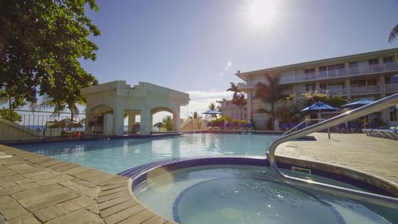 An outdoor pool reflecting sunlight, with sunbeds and sun huts at Courtleigh Hotel & Suites