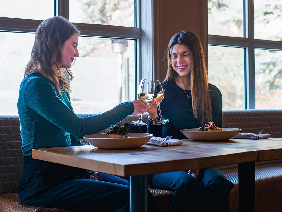 Two guests sitting at a table in a restaurant, clinking wine glasses, and smiling.