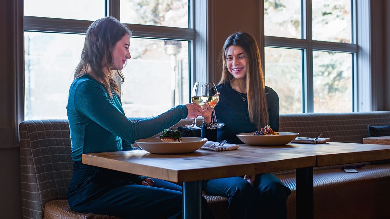 Two guests sitting at a table in a restaurant, clinking wine glasses, and smiling.