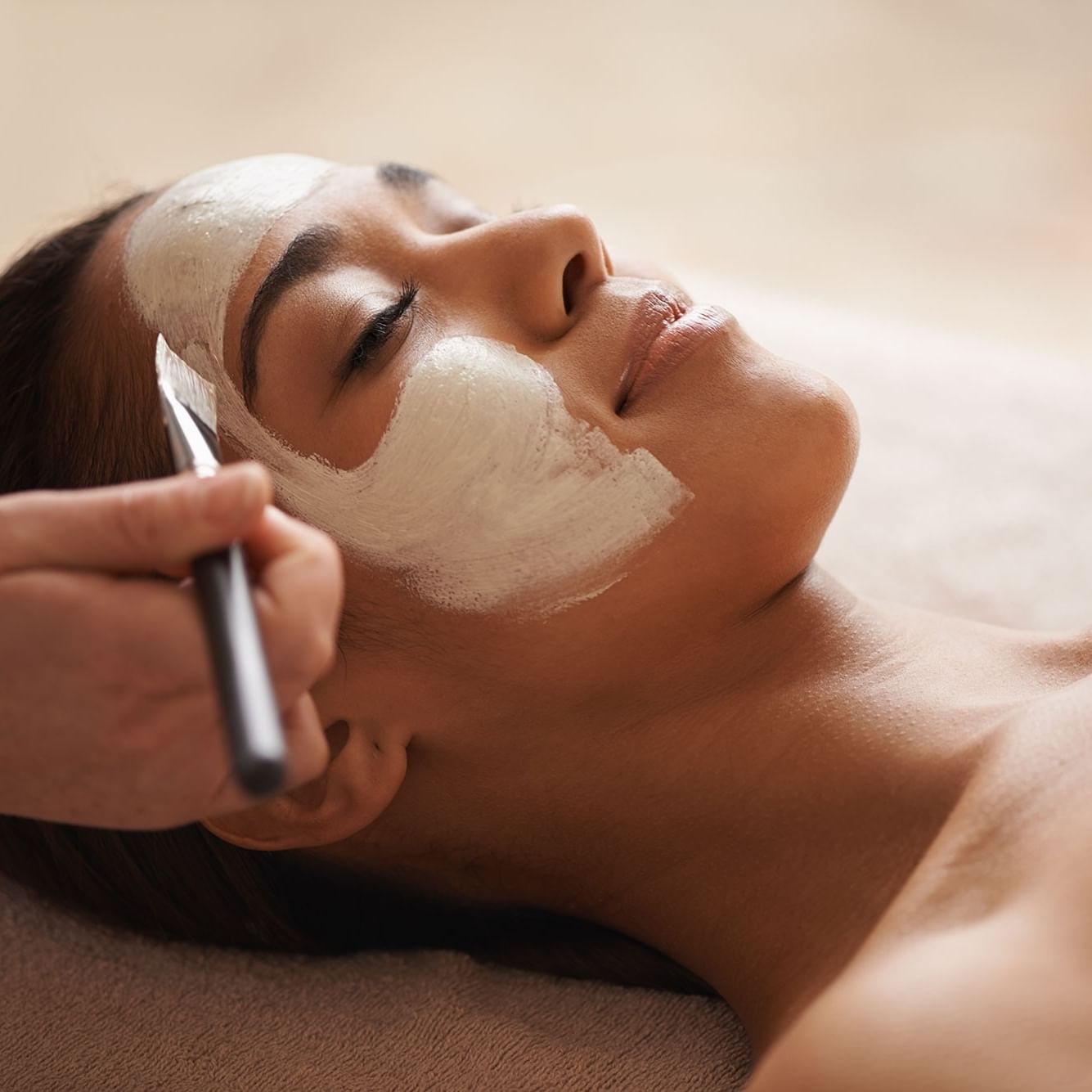 Woman receiving a facial treatment with a white mask being applied by a hand holding a tool.