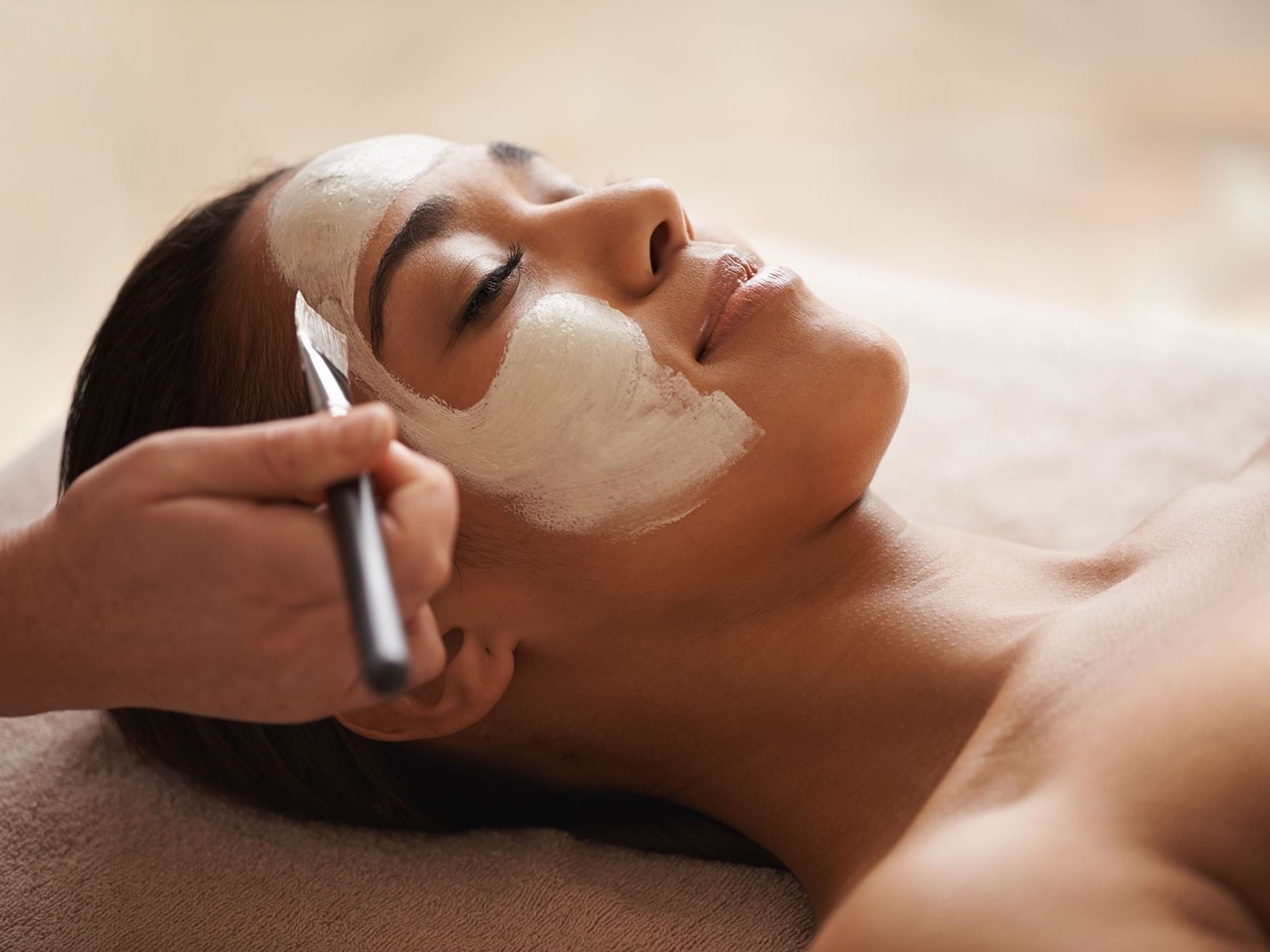 Woman lying down receiving a facial treatment with a mask and brush.