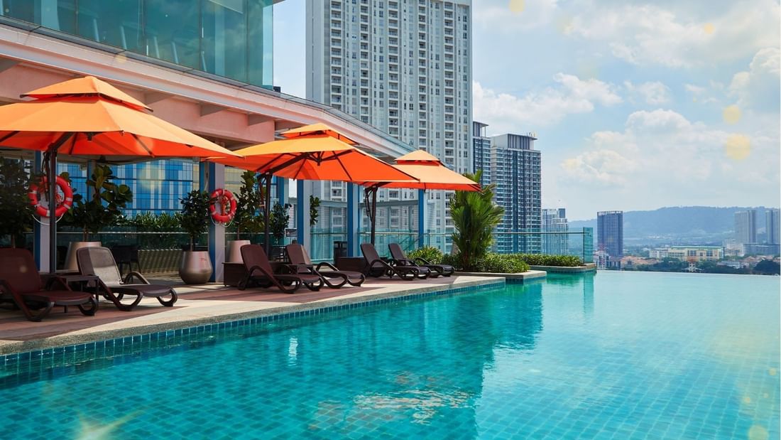 Outdoor pool with lounge chairs and umbrellas, surrounded by buildings under a partly cloudy sky.