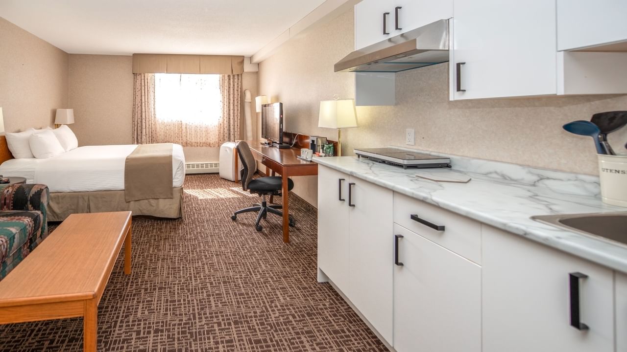 A hotel room with a bed, desk, chair, wooden table, and a kitchen with white cabinets.