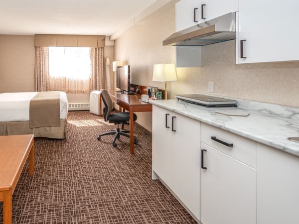 Hotel room with bed, desk, chair, TV, and kitchenette with sink and cabinets.