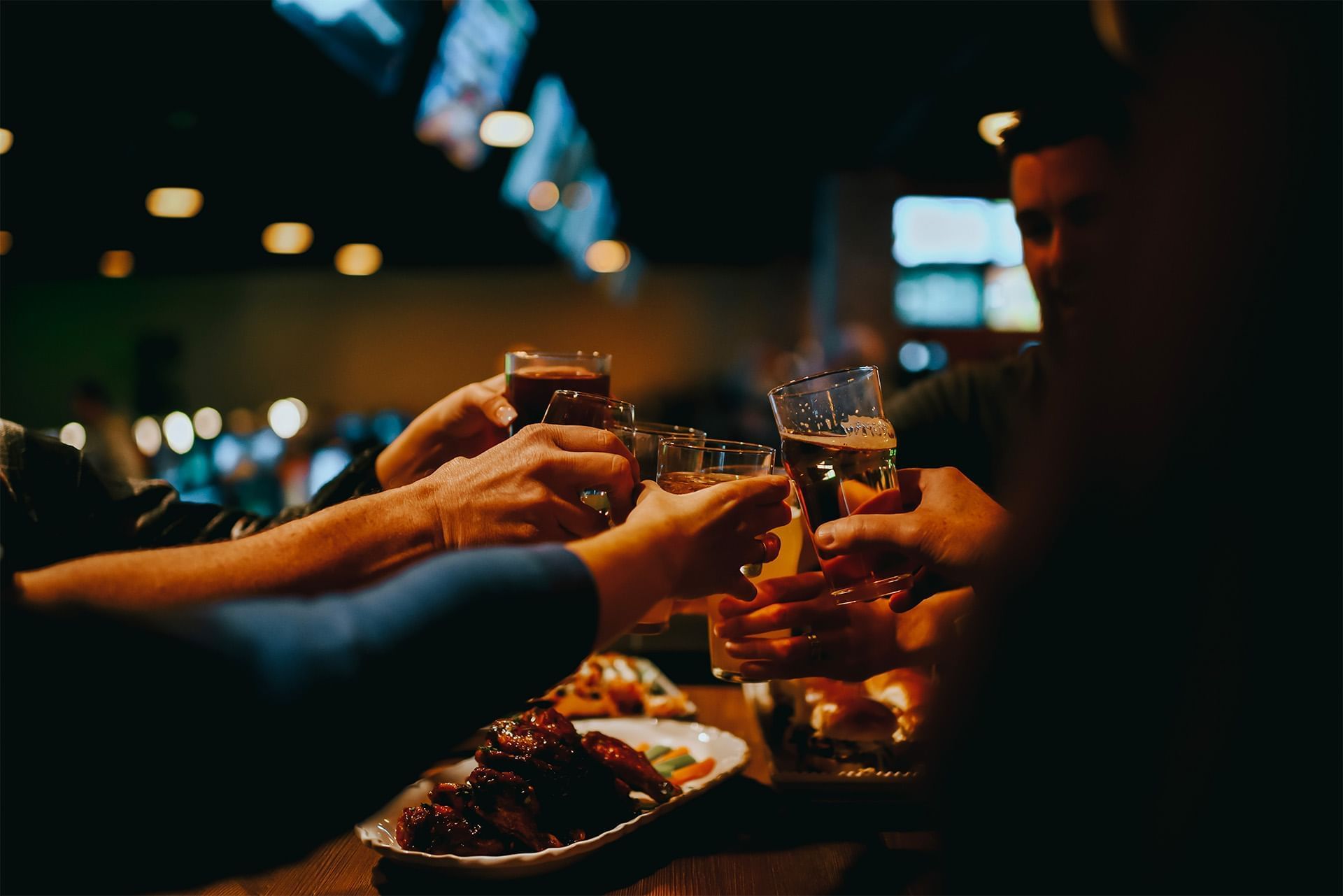 Friends raising beer glasses over plates of barbecue food under warm restaurant lights during a gathering at The Brook
