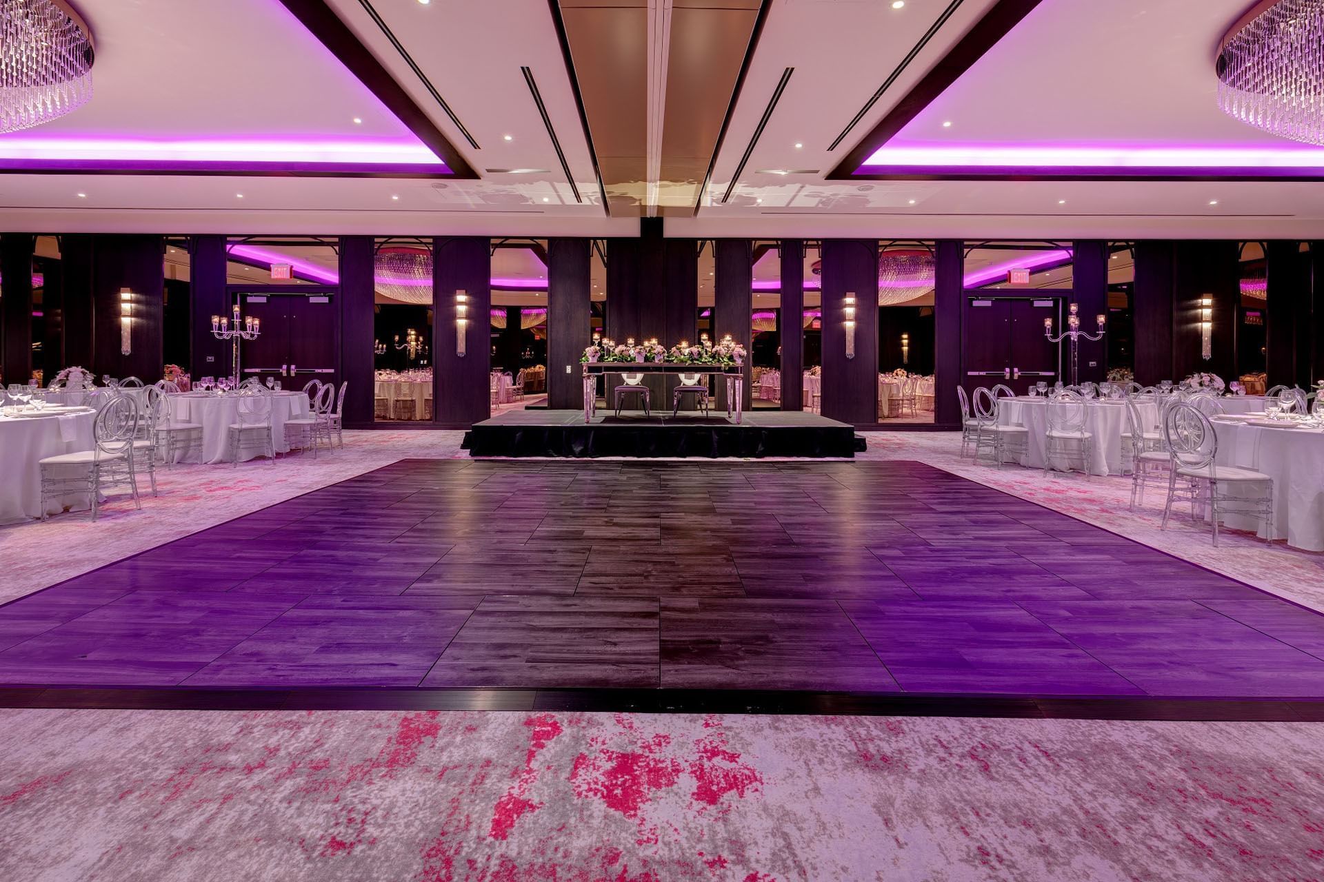 Princes Gates Ballroom at Hotel X Toronto with accent lighting and round tables & dark wooden dance floor near a small stage