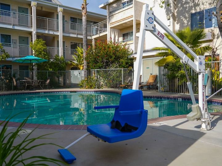 A blue pool lift chair sits by the edge of a swimming pool, with a white frame and buildings in the background.
