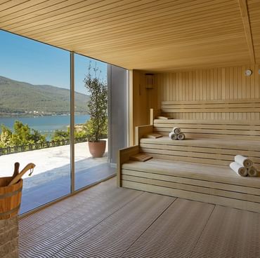 Деревянная сауна с стеклянными стенами, свернутыми полотенцами и видом на море и зелень — природное спокойствие и тепло в атмосфере Titanic Luxury Collection Bodrum.