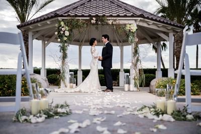 Bride and groom stand together in a flower-adorned gazebo with scattered rose petals at London Bridge Resort