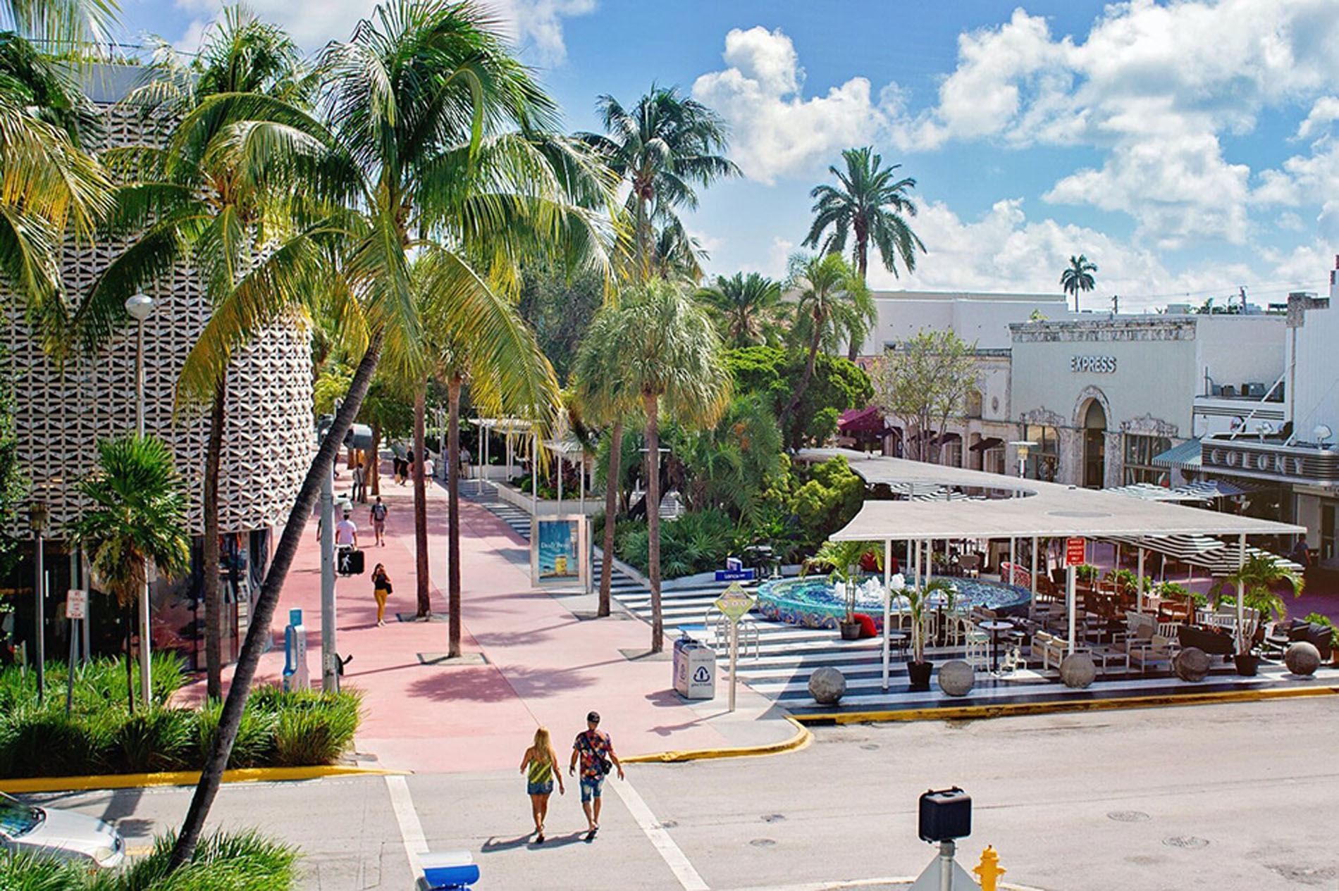 A street scene with palm trees, people walking, and buildings in the background.