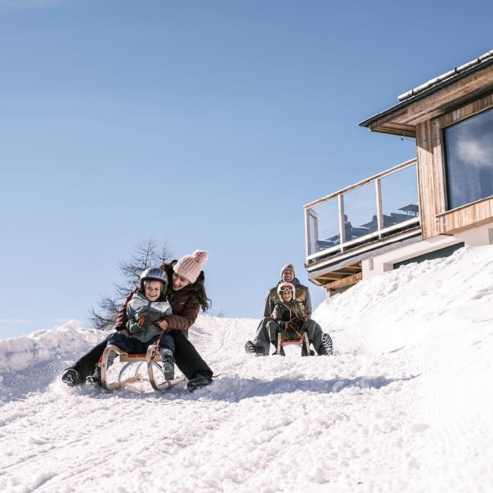 Famiglia su slittino in discesa innevata vicino a una casa di legno con terrazza.