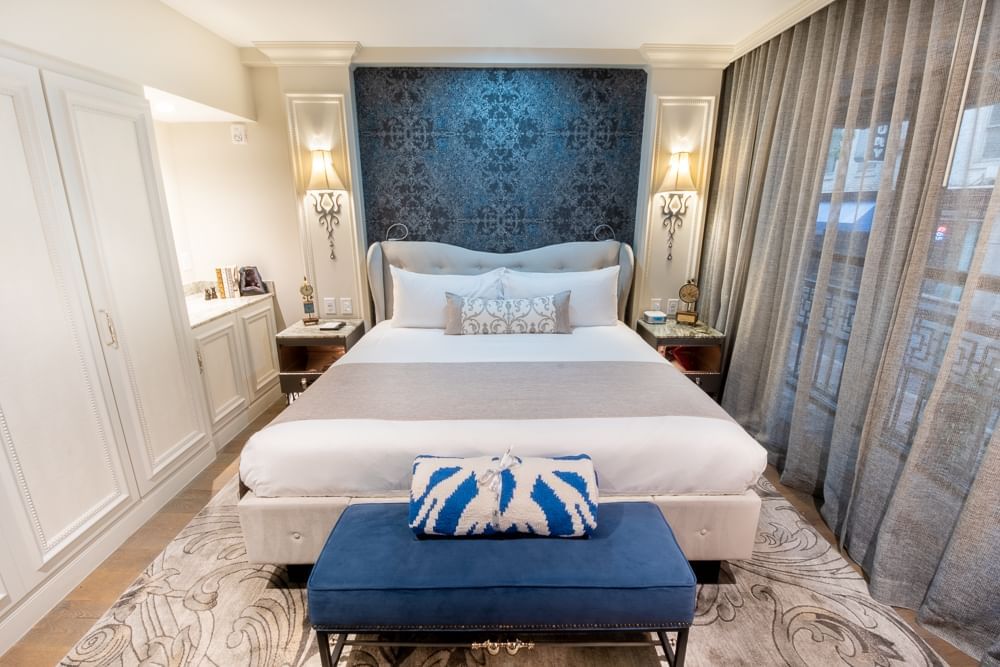 A hotel room with a large bed, blue bench, and gray curtains