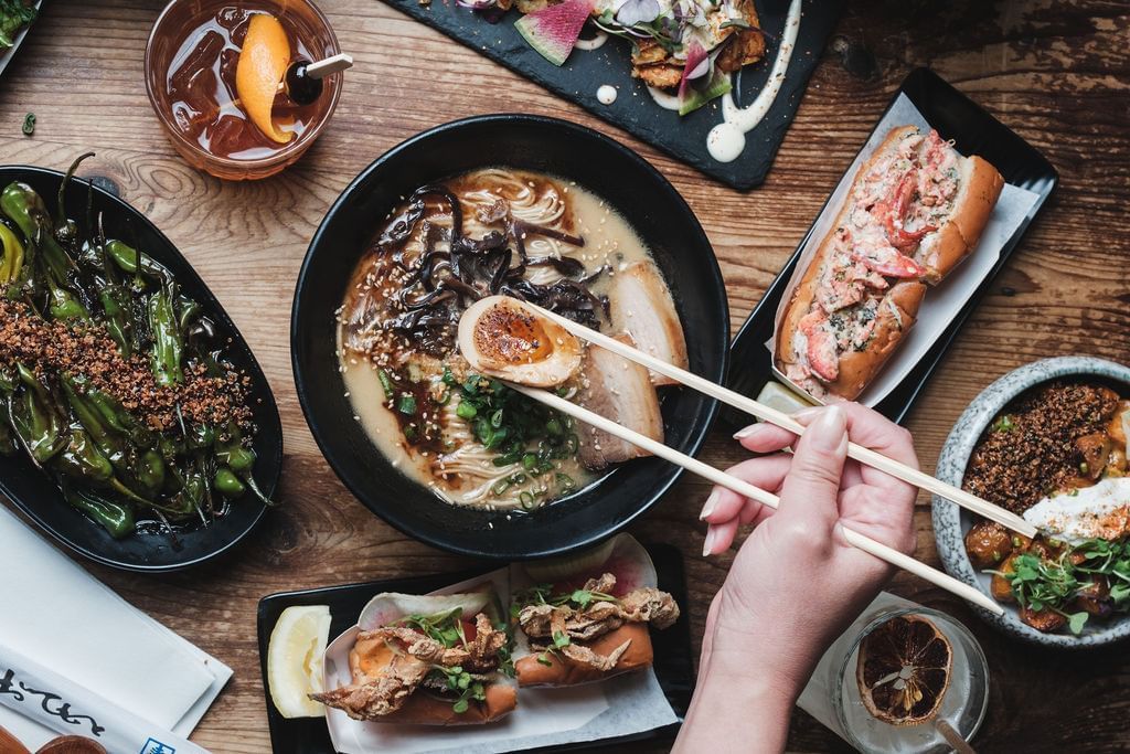 A photo from above of a hand holding chopsticks over a bowl of ramen surrounded by other dishes. 