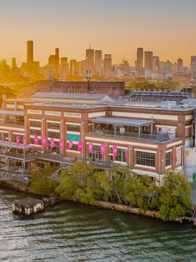 Aerial view of Brisbane Powerhouse arts center on the riverbank at sunset with city skyline in the background.