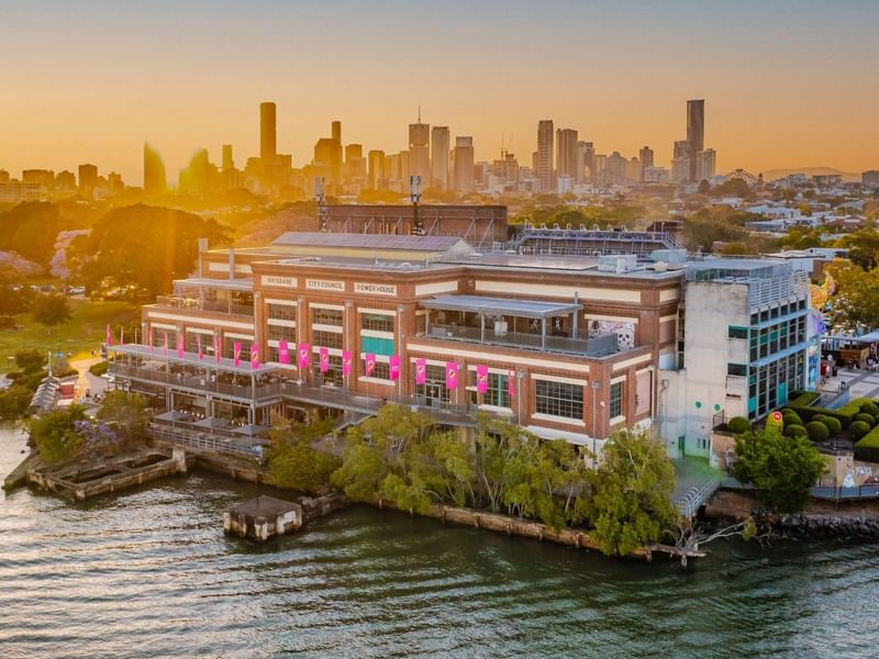 Aerial view of Brisbane Powerhouse arts center on the riverbank at sunset with city skyline in the background.