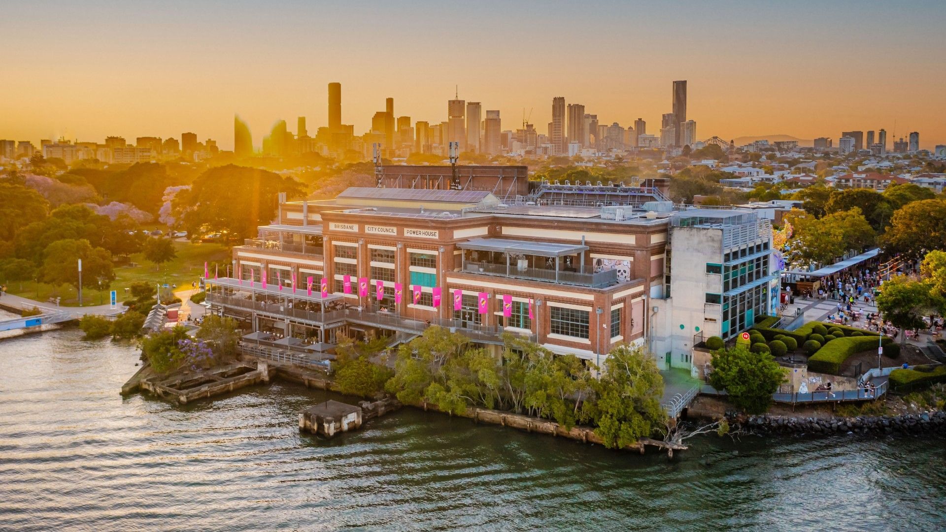 Aerial view of Brisbane Powerhouse arts center on the riverbank at sunset with city skyline in the background.
