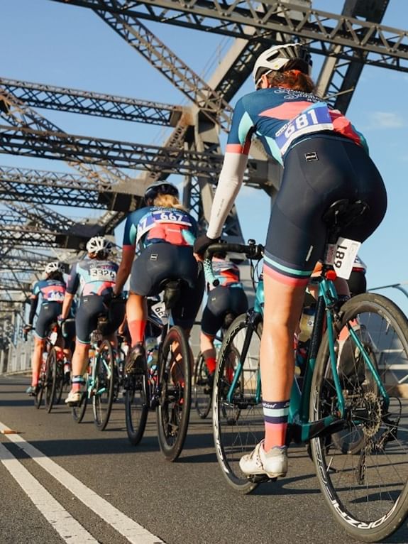 Cyclists race on a bridge with a large metal structure above during the Tour de Brisbane.