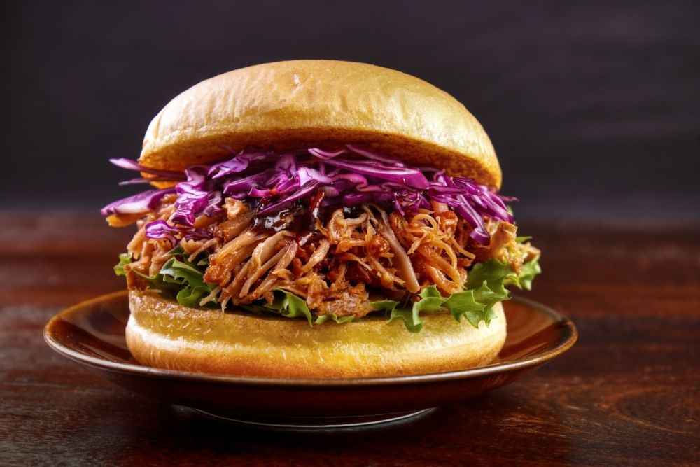 A sandwich on a small brown plate made up of a bun and layers of purple cabbage, shredded barbecue meat, and lettuce.
