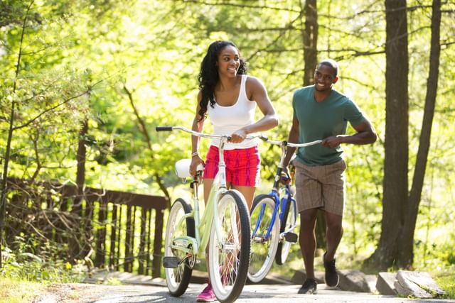 {Two individuals walk their bicycles along a sunny forest trail near Cove Pocono Resorts}