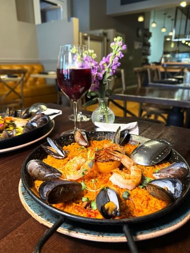 Seafood paella with a pan by a glass of red wine on a wooden table in BOTA at Warwick San Francisco