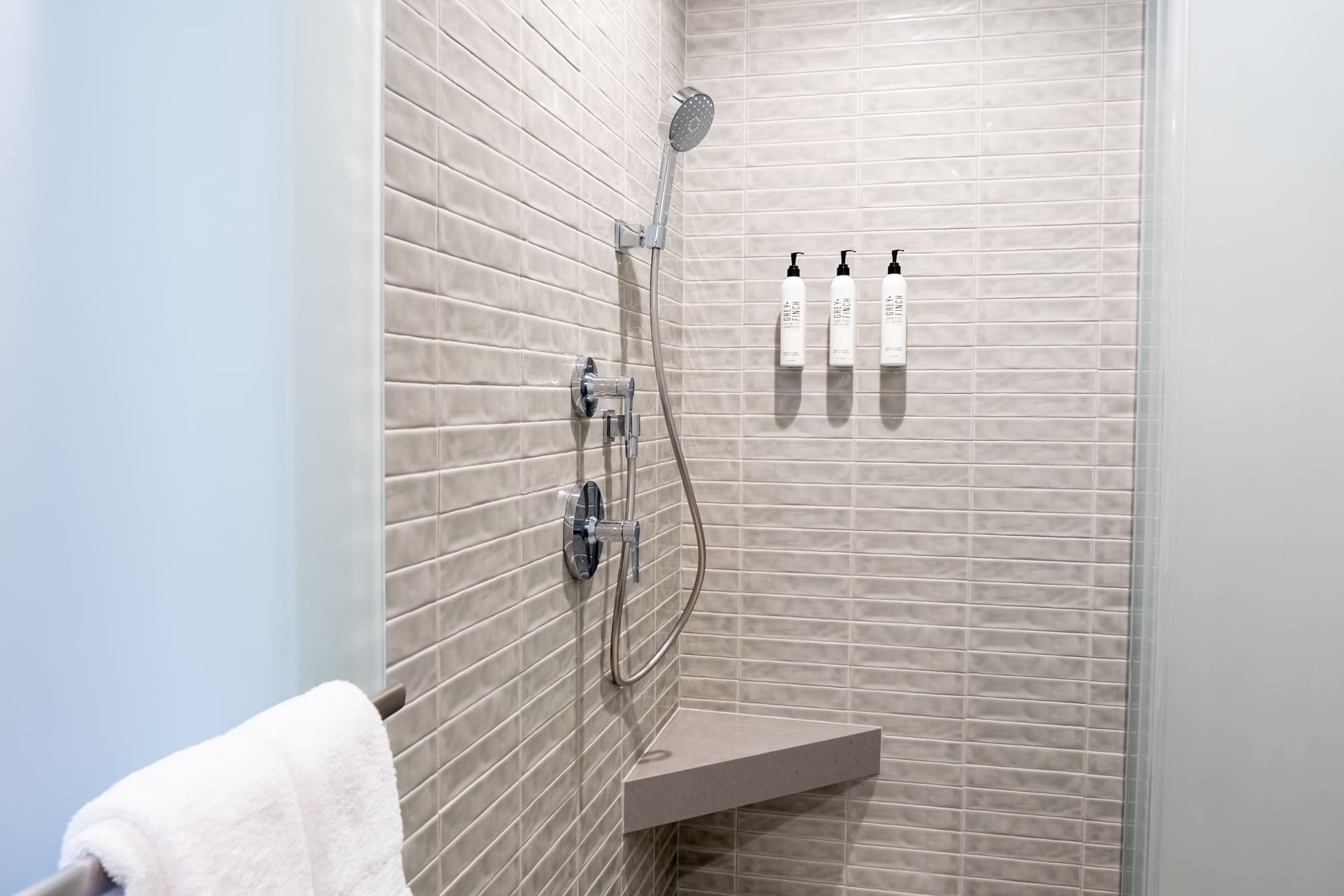 A modern shower with tiled walls, a shower head, and a shelf with towels and soap bottles.