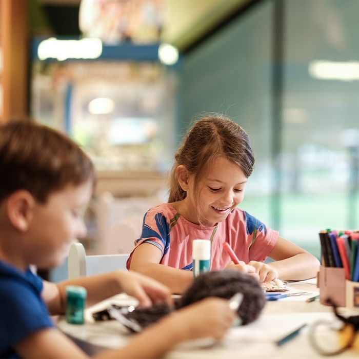 Due bambini lavorano insieme su un progetto artigianale con colla, forbici e pennarelli.