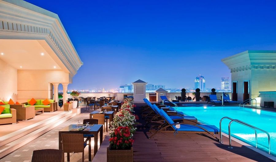Evening view of rooftop pool area with lounge chairs and dining tables at Warwick Doha Hotel