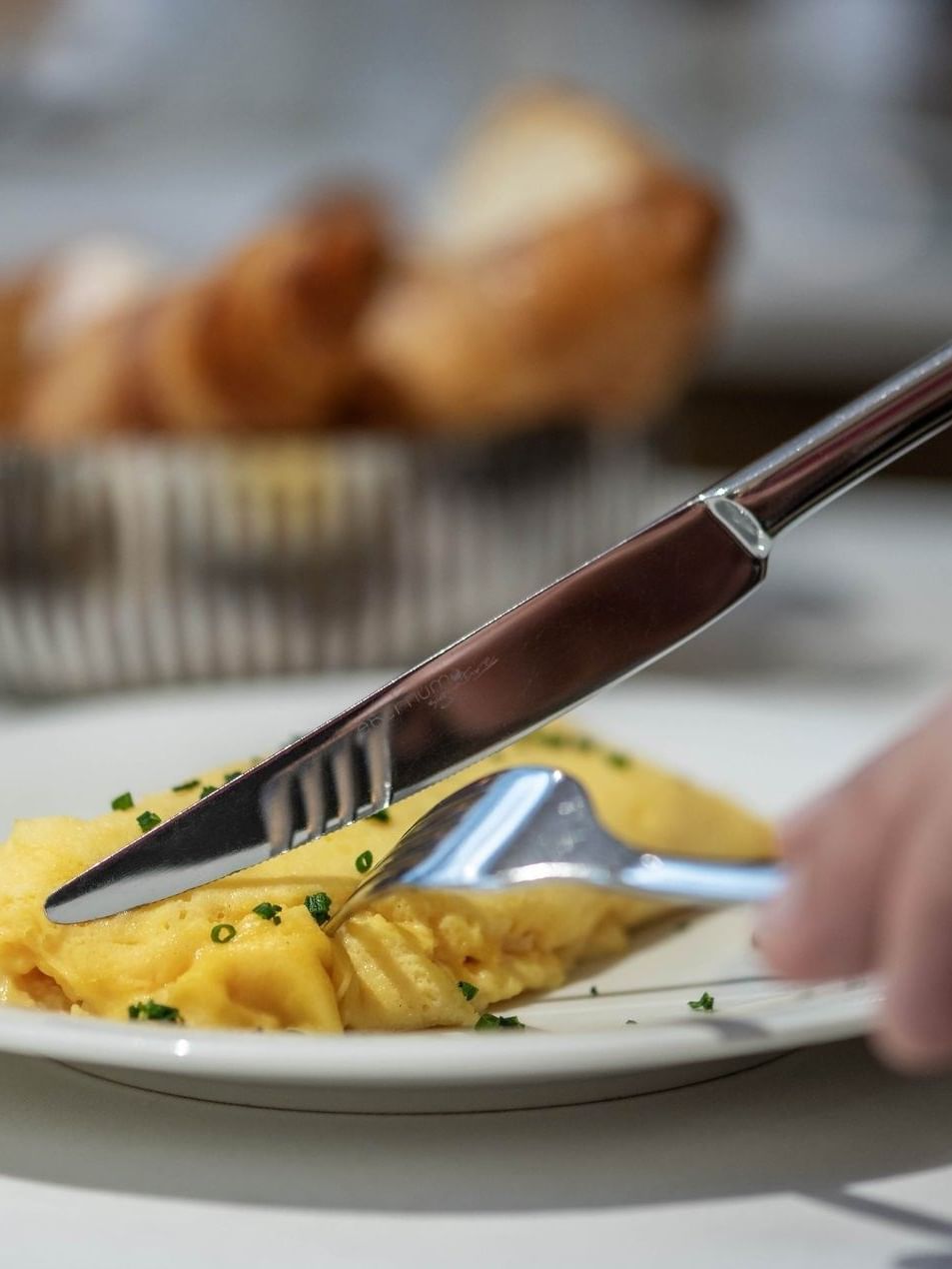 Close-up of a guest using a silver knife and fork to cut a chive-topped omelet by a bread basket at Hotel Westminster Paris
