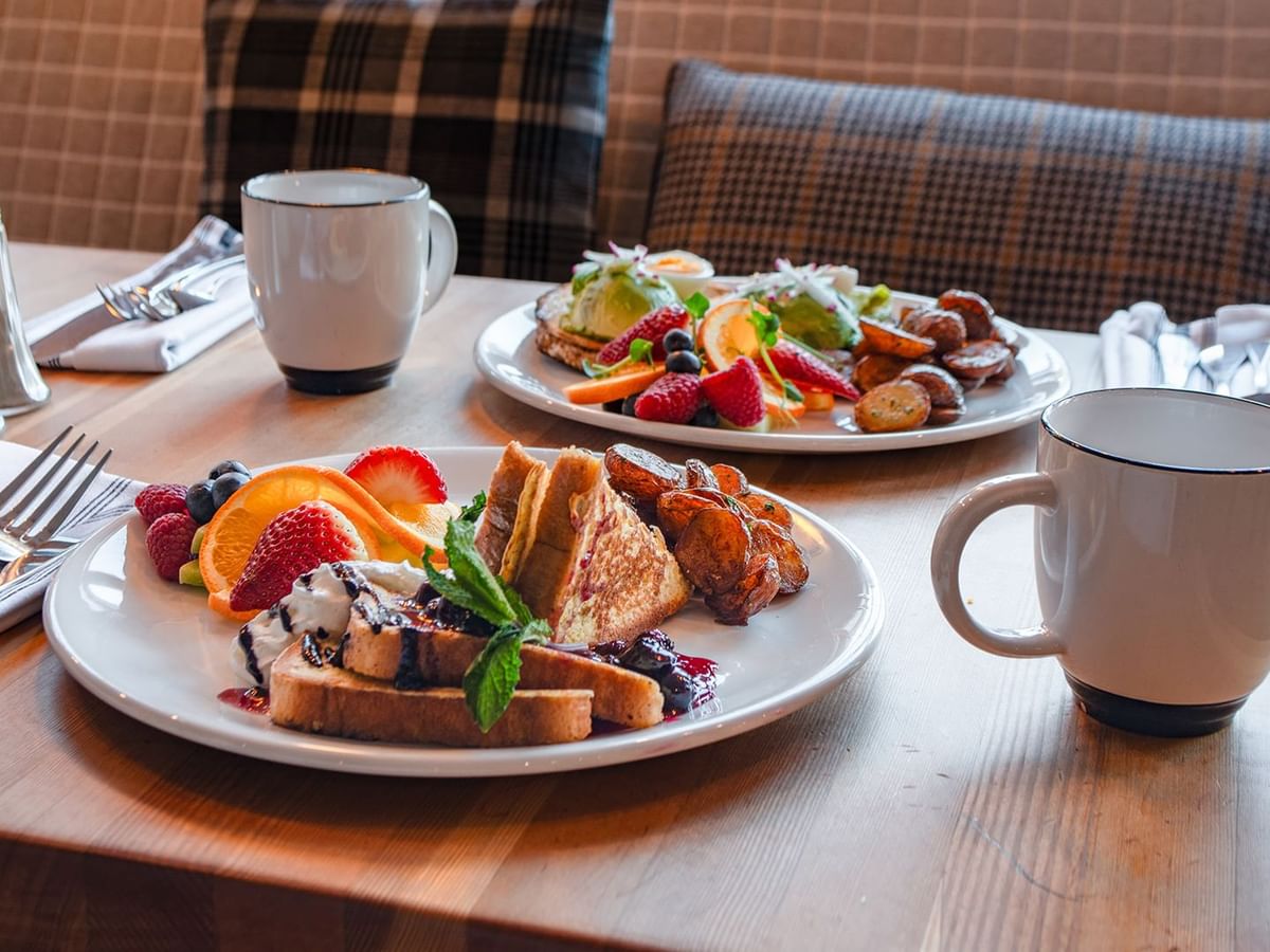 Two plates of food with fruits and pastries, accompanied by coffee mugs and cutlery on a table.