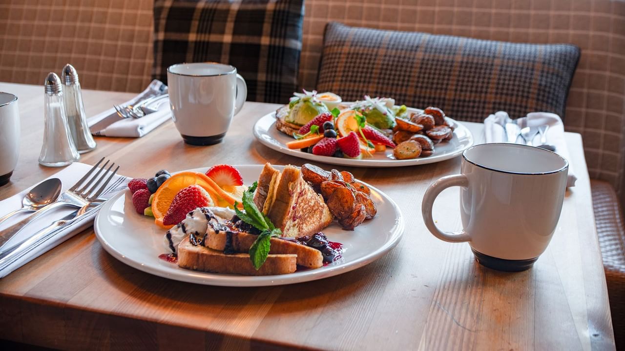 Two plates of food with fruits and pastries, accompanied by coffee mugs and cutlery on a table.