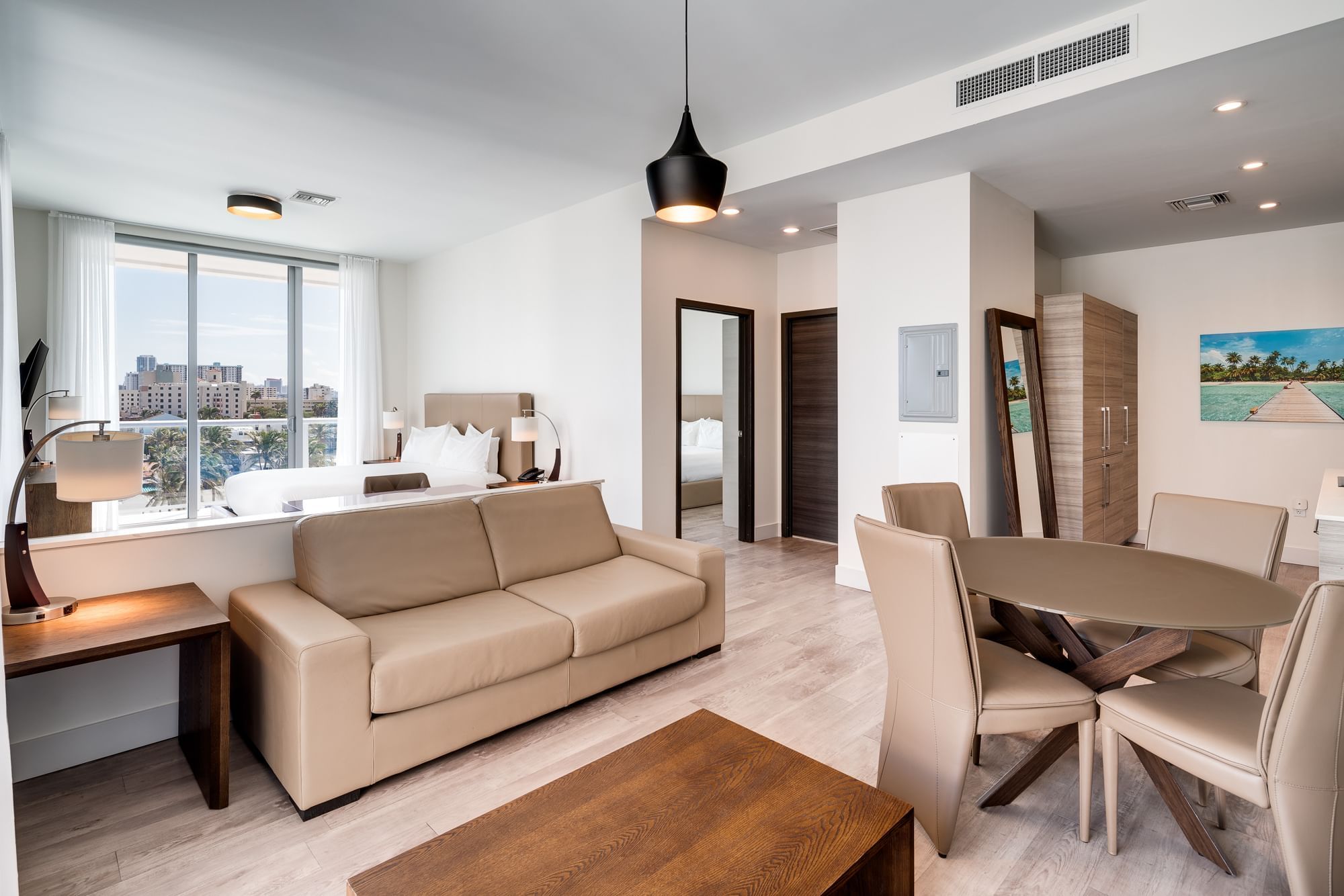 A wide view of a hotel living room with a sofa, dining table, and city view.