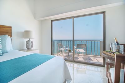 An elegant resort bedroom layout looking out through sliding glass doors to a sunny oceanfront balcony.