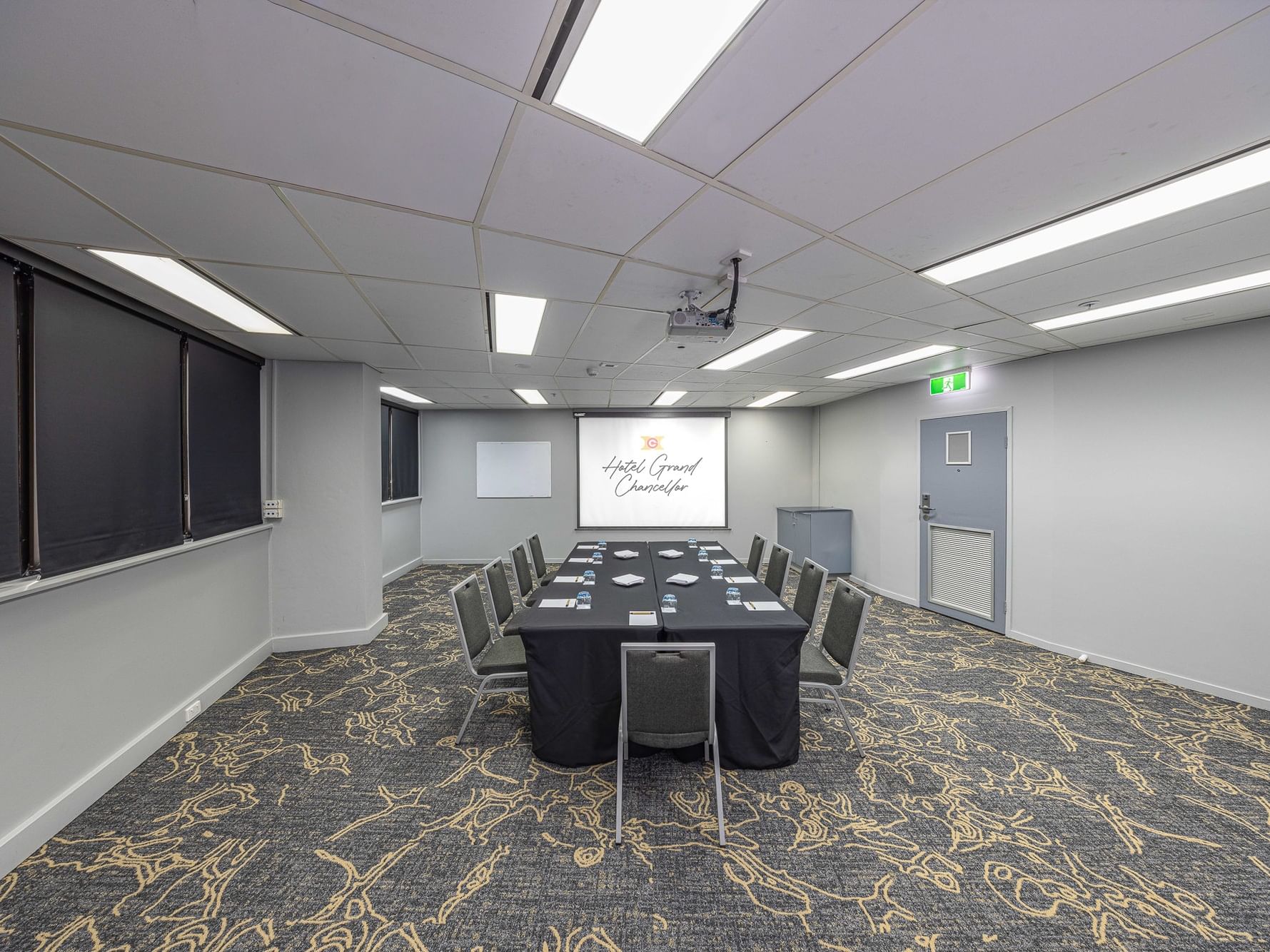 Professional hotel function room configured for a board meeting, featuring patterned carpet, privacy blinds, and a presentation setup.