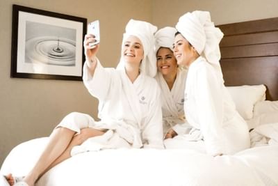 Three women in bathrobes smiling and posing for a selfie on a bed at Temple Gardens Hotel & Spa