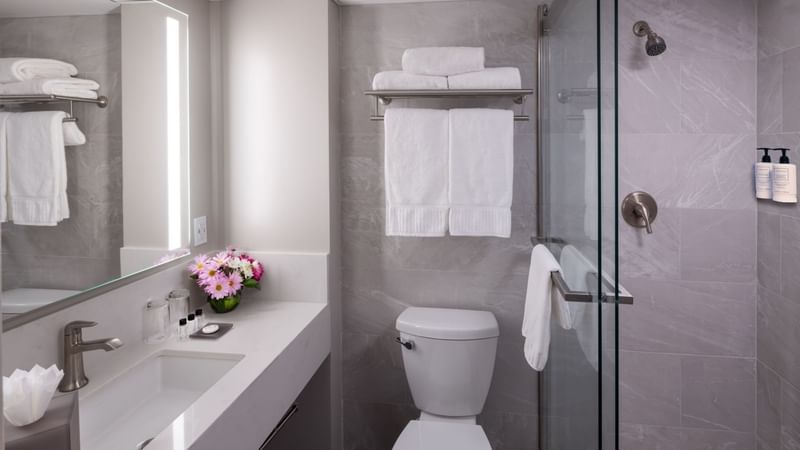 Modern bathroom with white toilet, sink, mirror, and glass shower with towels and flowers at Warwick Hotels and Resorts