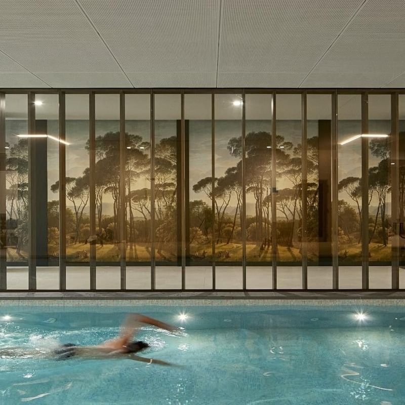Person swimming in an indoor pool with a forest mural on the wall and vertical blinds.