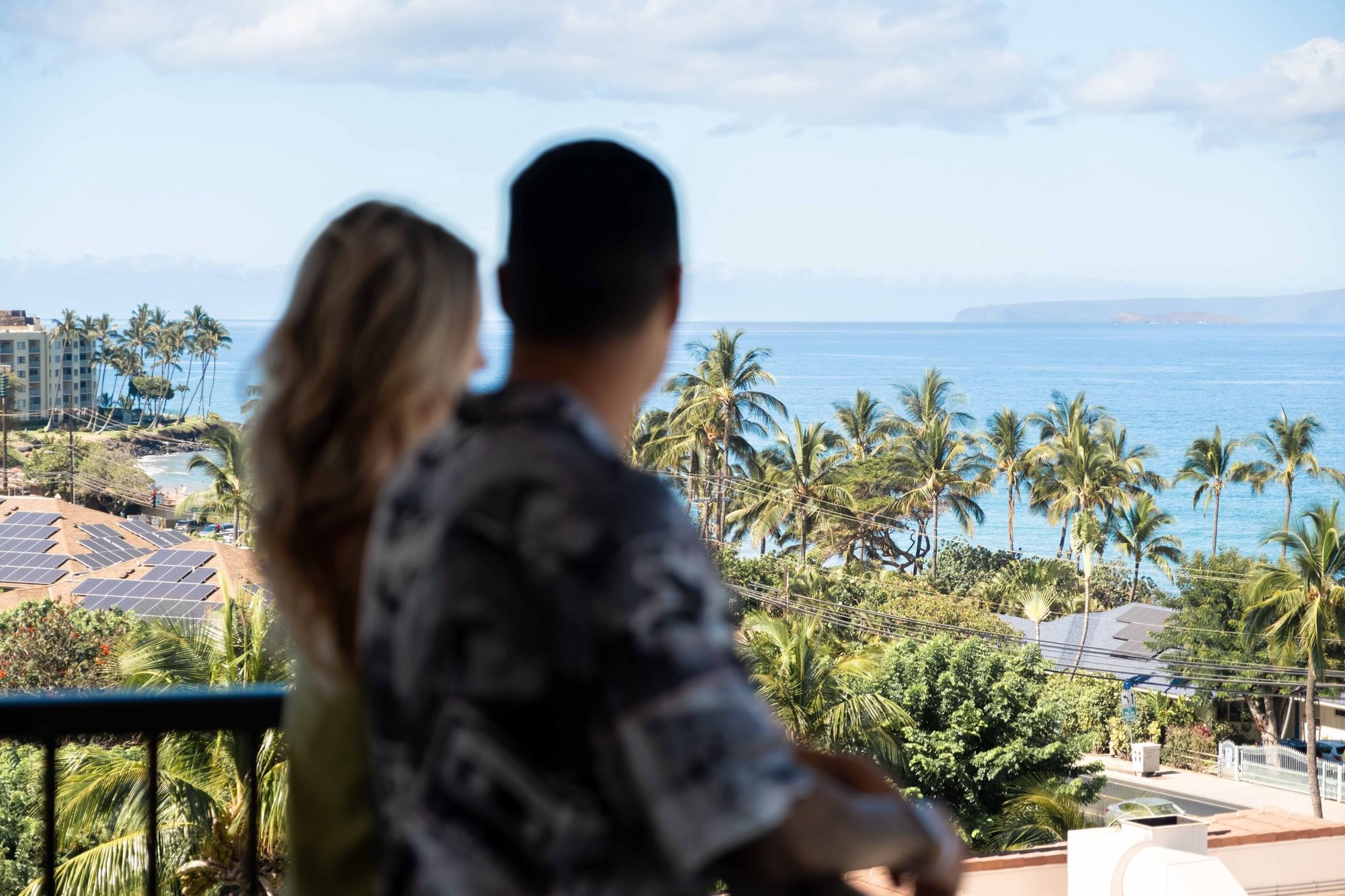 A couple on a balcony overlooking the ocean with palm trees and a tropical landscape.