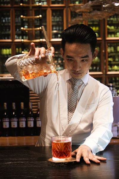 Bartender pouring a dark drink over ice from a textured glass pitcher in Opera Bar at Park Hyatt Saigon