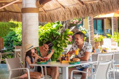 Family dining outdoors under a thatched roof with tropical decor and vibrant greenery in the background.