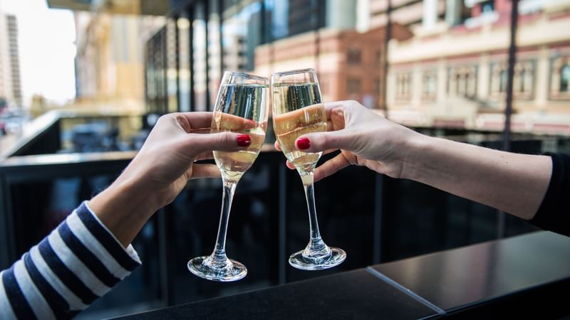 Two people clinking champagne glasses together in a toast, with a city view behind at Ibis Adelaide