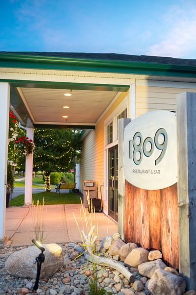 Restaurant sign by a stone garden bed under an outdoor awning, with trees in the yard at Rodd Miramichi River