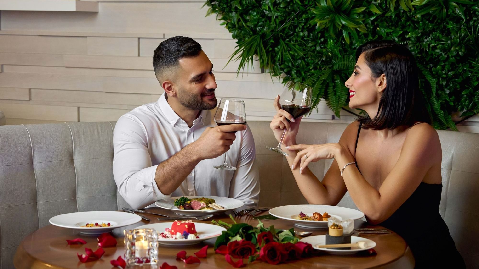 A happy couple enjoying a romantic dinner with red wine and spreading rose petals on the table at Paramount Hotel Dubai