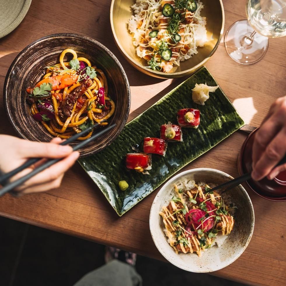 Overhead view of assorted Asian fusion dishes including noodles, sushi rolls, and garnished bowls on a wooden table.