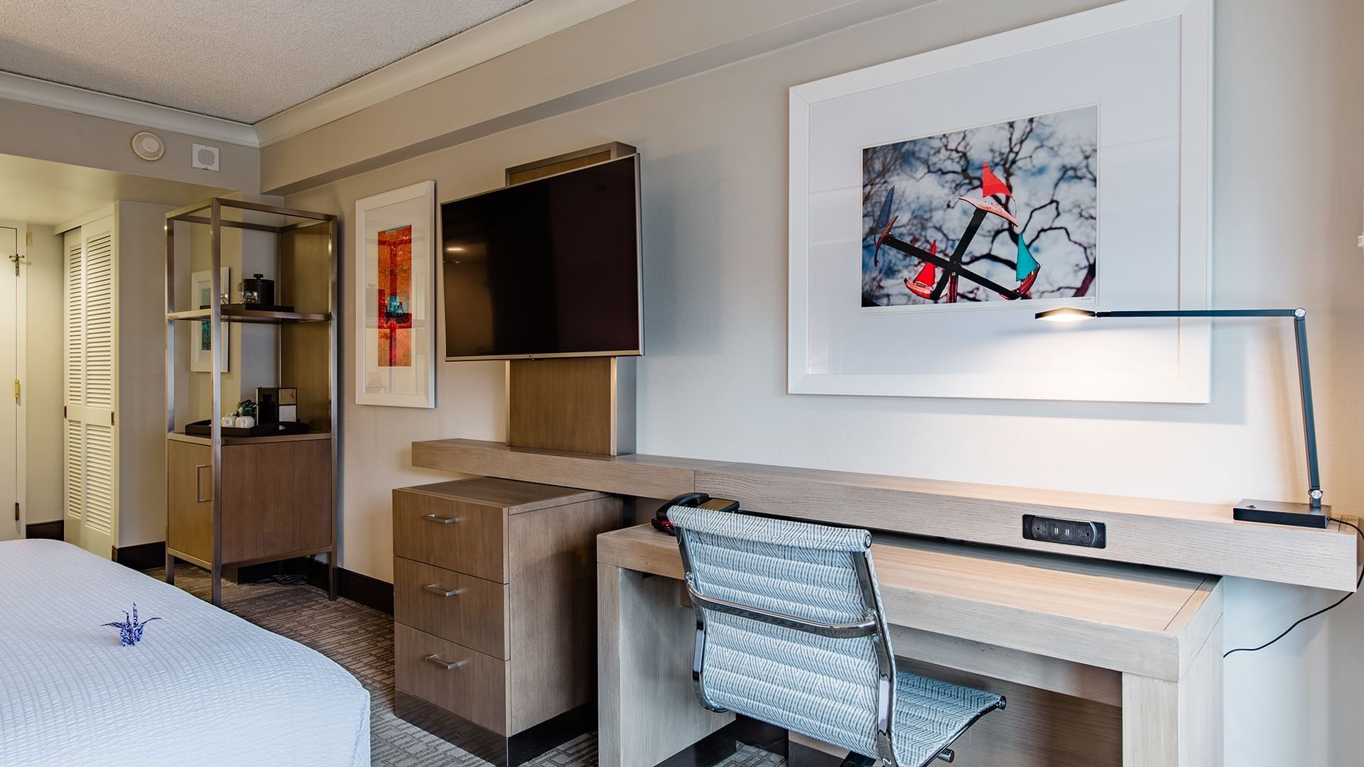 A hotel room with a desk, chair, TV, and artwork