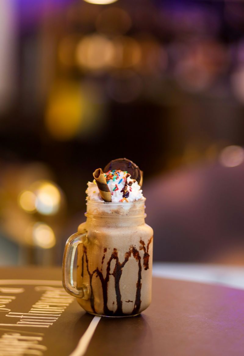 A chocolate milkshake in a glass jar, topped with whipped cream, a cookie, a rolled wafer, and colorful sprinkles, with chocolate syrup drizzled down the sides of the jar.