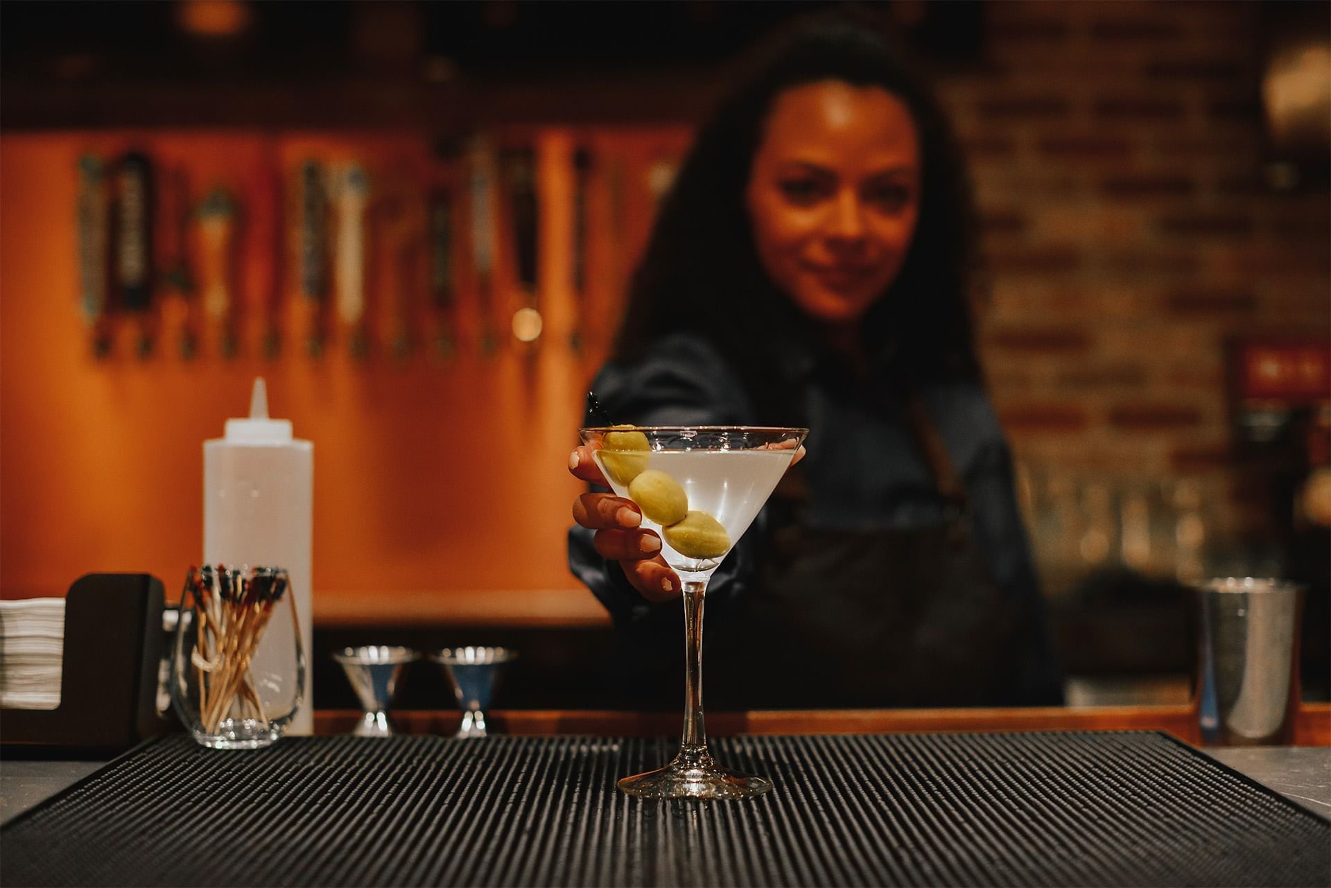 Bartender handing over a chilled martini with olives across a bar counter by cocktail shakers and skewers at The Brook